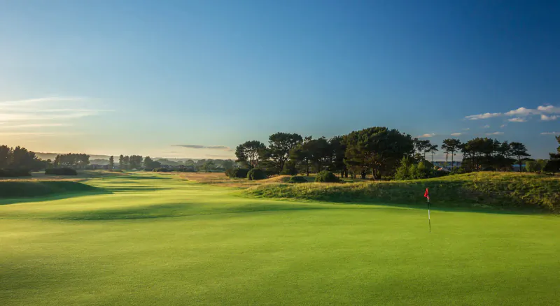 Carnoustie Links golf course at sunset, lush green fairway with bunkers, trees, and red flag on horizon.