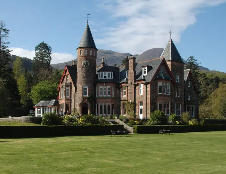 Red sandstone turreted mansion with conical towers amid Scottish mountains, gardens, and lawns under blue sky.
