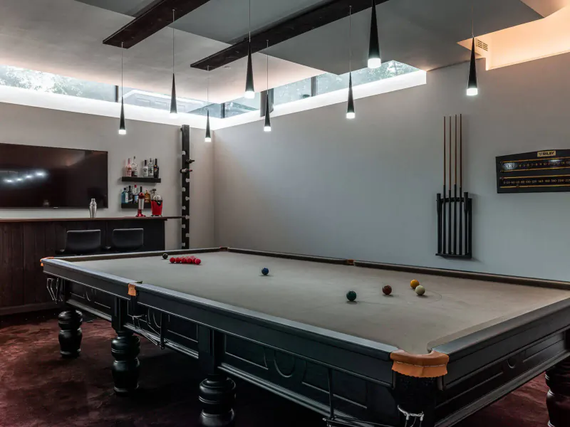 Modern glasshouse game room with black pool table, balls, cues, bar, and TV under skylight pendants.