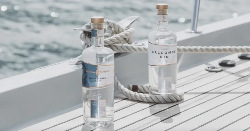 Two Salcombe Gin bottles on a yacht deck with ropes, cleats, and sparkling sea beyond.