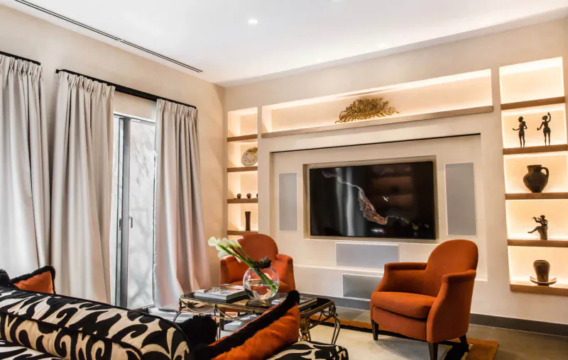 Elegant hotel living room with orange armchairs, black sofa, large TV, shelves with African statues, and flowers.