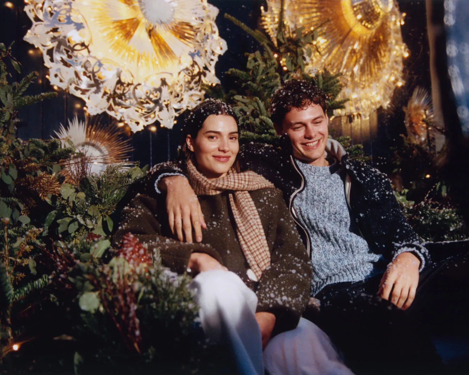 Smiling couple in coats and scarf, embracing amid festive gold wreaths and greenery at Bicester Village