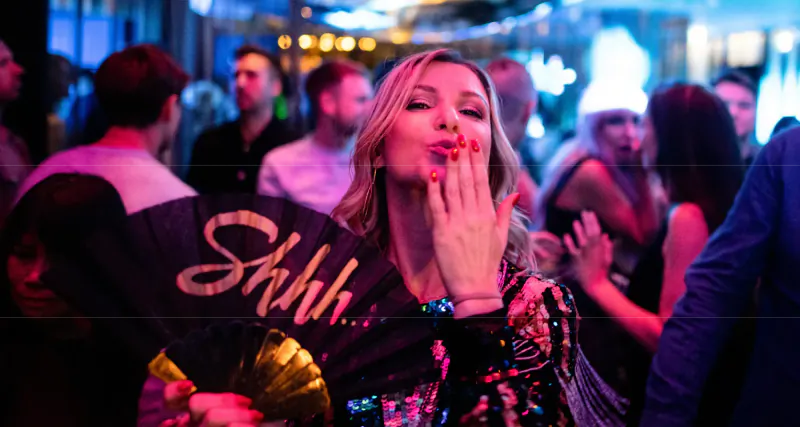 Blonde woman blowing kiss with hand, holding 'Shhh' fan, in vibrant neon-lit Le Mans party crowd