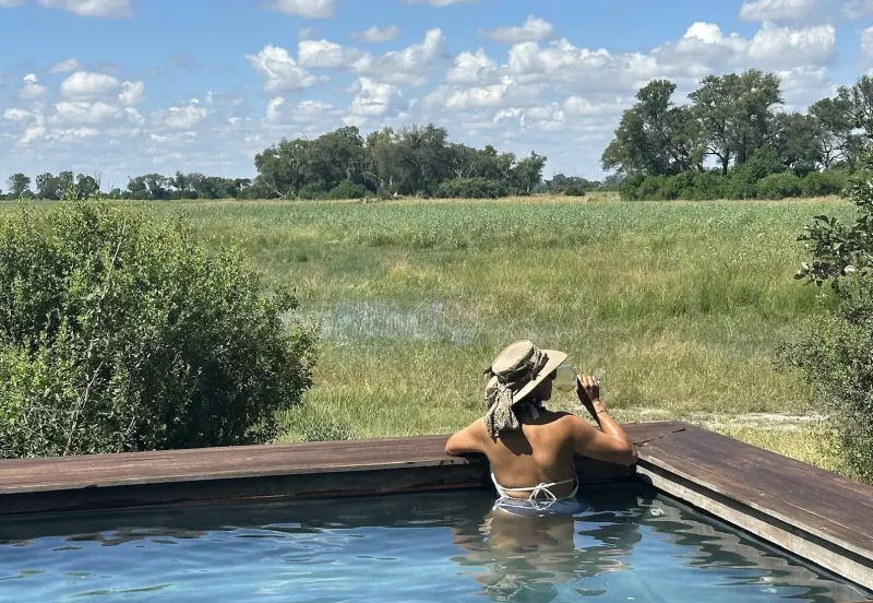 Woman in beige sunhat and bikini relaxes in pool, leaning on wooden edge, gazing at Botswana savanna with trees and wetlands.