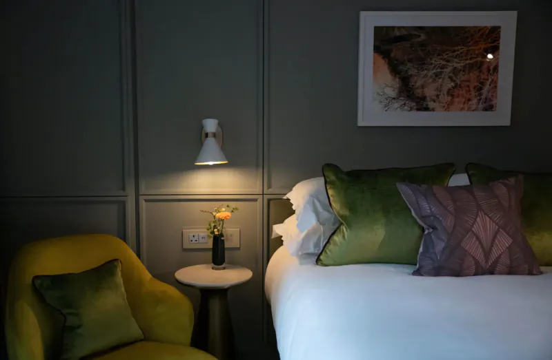 Cozy hotel bedroom with white bed, green and purple pillows, yellow armchair, vase of flowers, wall lamp, and rocky landscape art.