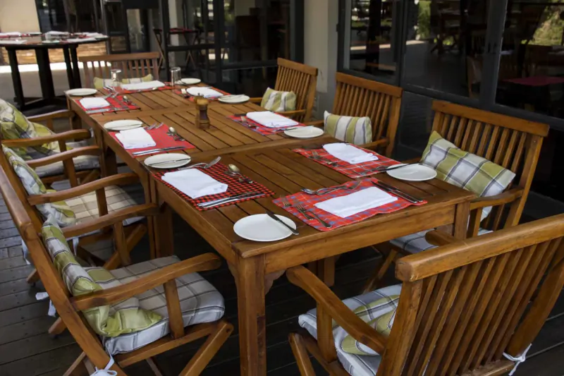 Elegant outdoor wooden dining table set for multiple guests with red plaid placemats, cushions on chairs, at Norfolk Nairobi.