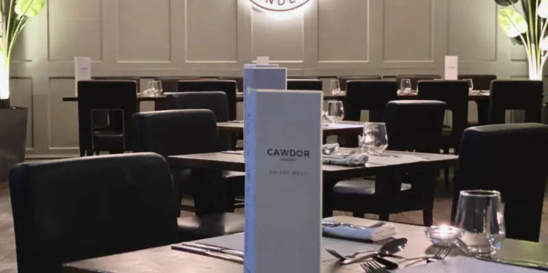 Elegant dining room at Cawdor Hotel with set tables, 'Cawdor Hotel' menu stands, and plants in soft lighting.