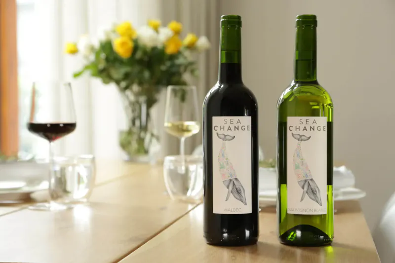 Two Sea Change wine bottles, red and white, with glasses of wine and yellow flowers on a table.