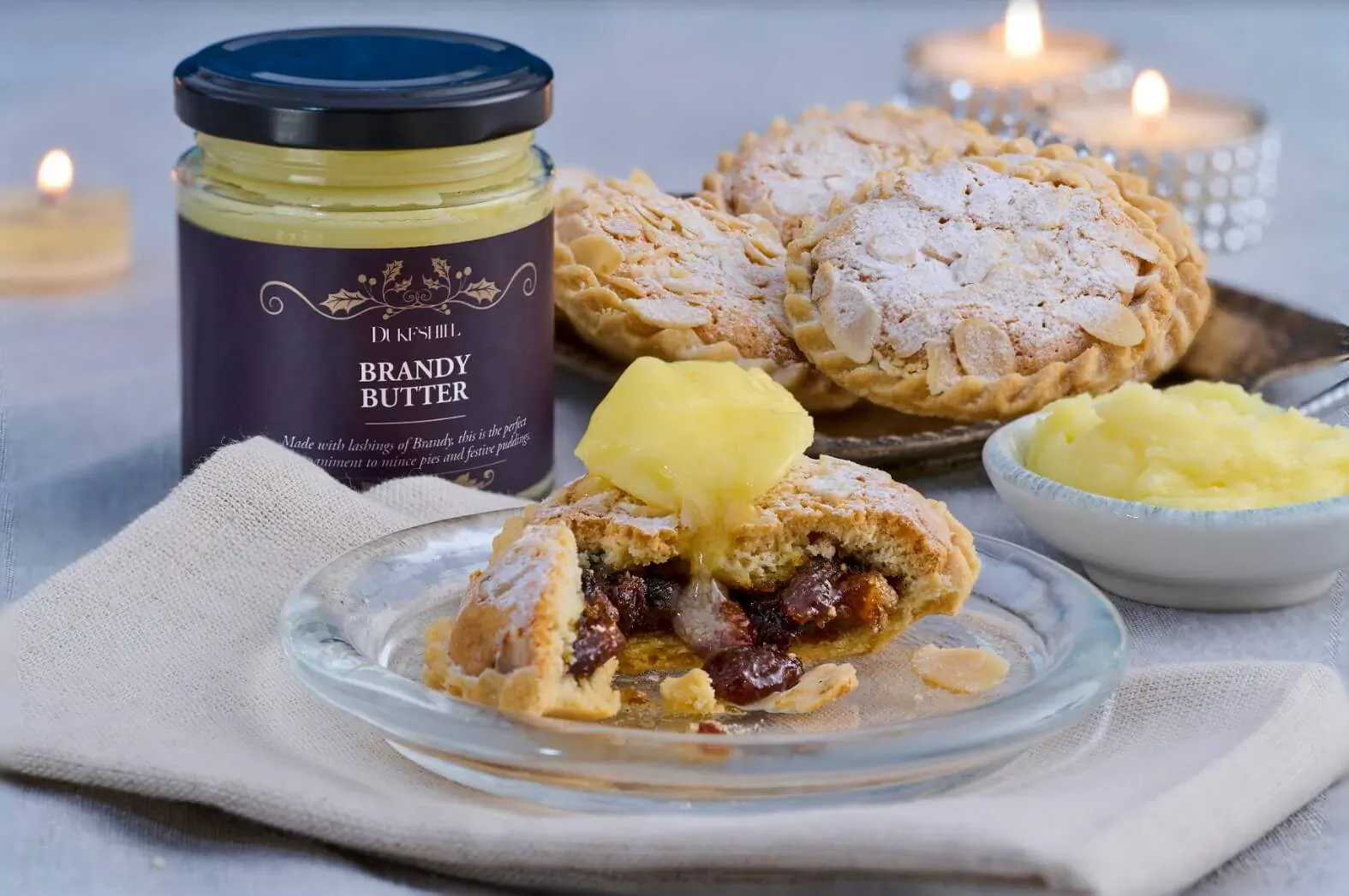 Dukeshill brandy butter jar beside open tart with brandy butter drizzle, raisins, almonds on plate, candles lit.
