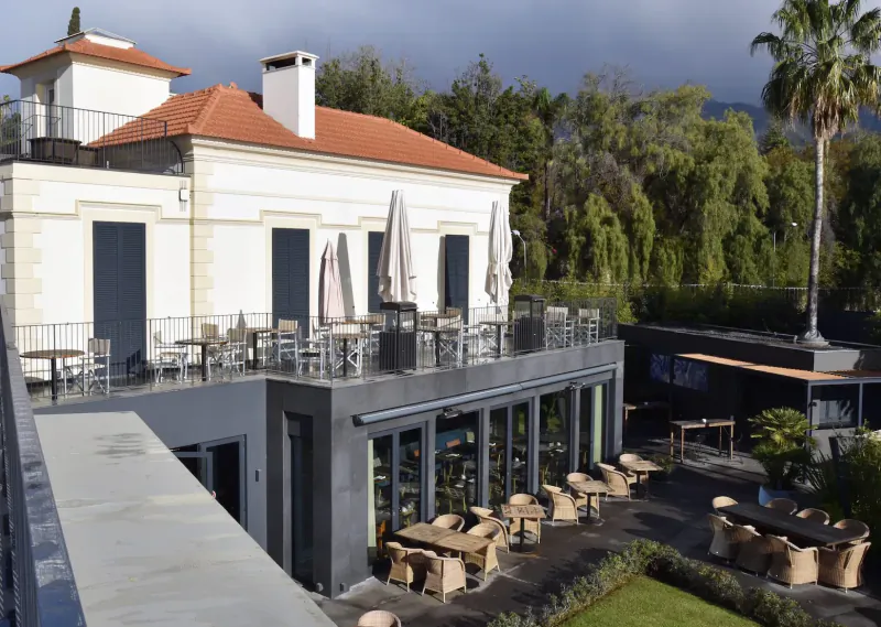 Les Suites Hotel at The Cliff Bay in Funchal, Madeira: white building with red roof, terrace with tables, chairs, umbrellas, garden, palms, mountains.