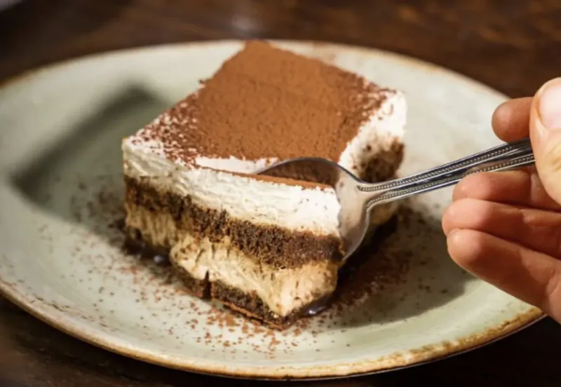Hand scooping tiramisu layered with mascarpone and cocoa from a white plate on wooden table.