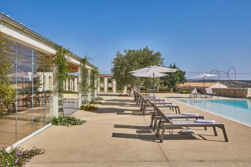 Luxury Sicilian hotel poolside with lounge chairs, umbrellas, olive trees, and modern glass pavilion under blue sky.