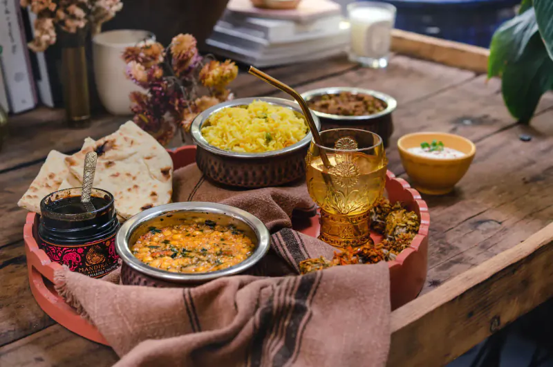 Indian thali on wooden tray: yellow rice, dal, curry, naan, raita, chutney, with flowers and milk glass.