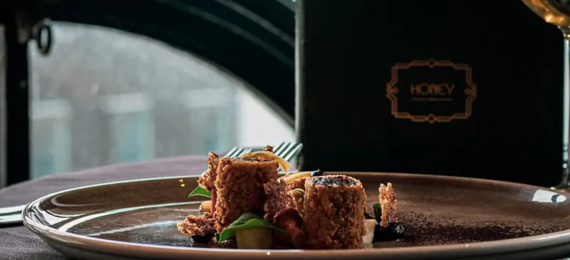 Three breaded cylindrical dishes with greens on a plate at Hotel Gotham restaurant, beside wine glass and menu.