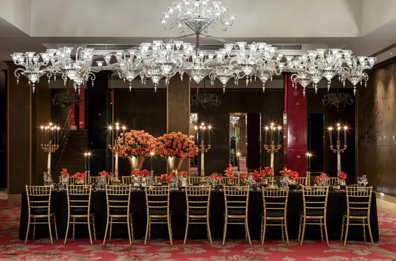 Elegant hotel banquet room with large crystal chandeliers, orange floral centerpieces, black chairs, and red carpet.