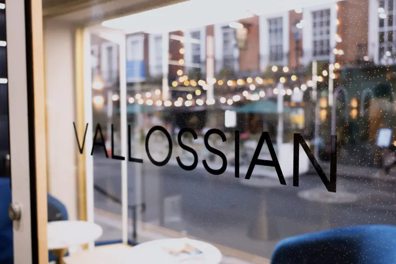 Vallossian storefront window with 'Vallossian' lettering, blue chairs inside, festive street lights outside at night