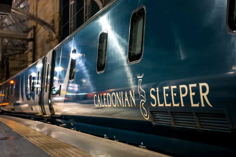 Caledonian Sleeper train at night station platform, illuminated with 'Caledonian Sleeper' text on side.
