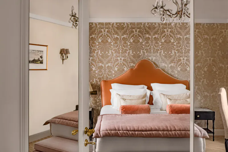 Luxurious bedroom with orange tufted headboard, beige pillows, ornate wallpaper, chandelier, and mirror reflection at Relais Christine Paris.