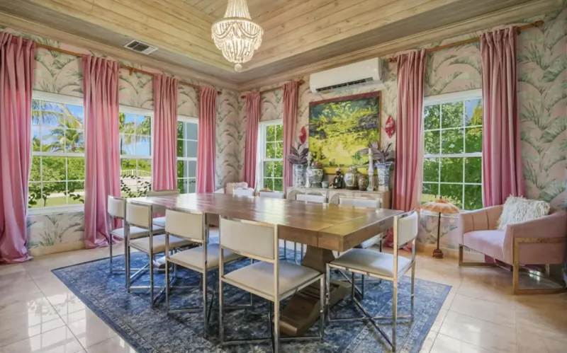 Elegant dining room in Lyford Cay villa with wooden table, pink floral wallpaper, chandelier, abstract painting, and garden views.