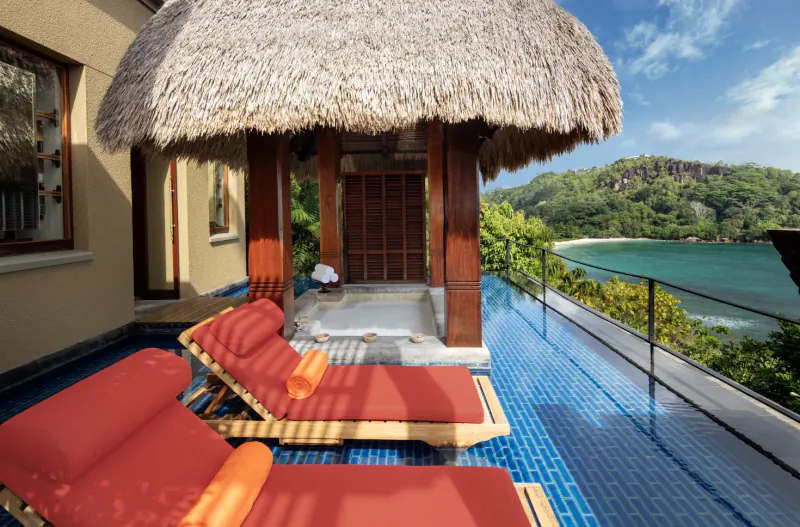 Luxury thatched villa deck with red loungers and blue tiles overlooking Seychelles beach and hills