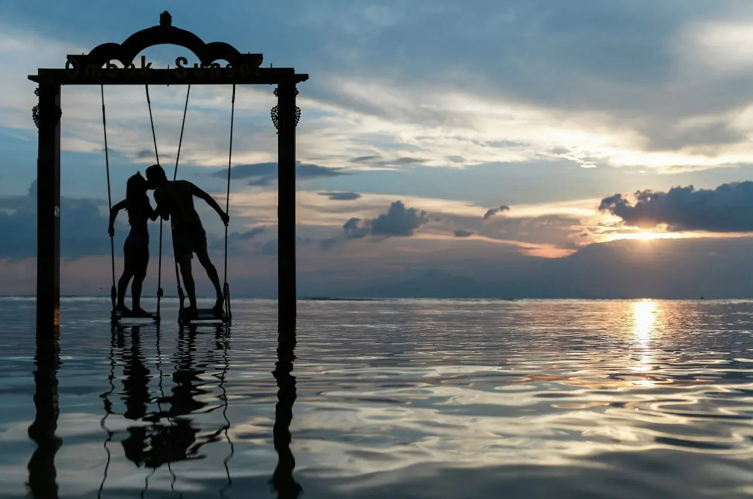 Silhouette of couple kissing on swing in ornate frame, reflected in sunset ocean water.