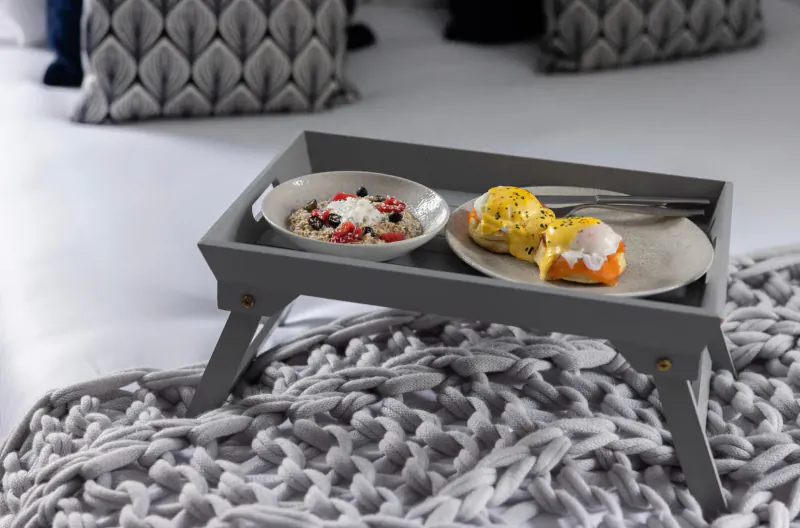 Grey wooden lap tray on knitted blanket holding yogurt parfait with berries and fried egg with hollandaise