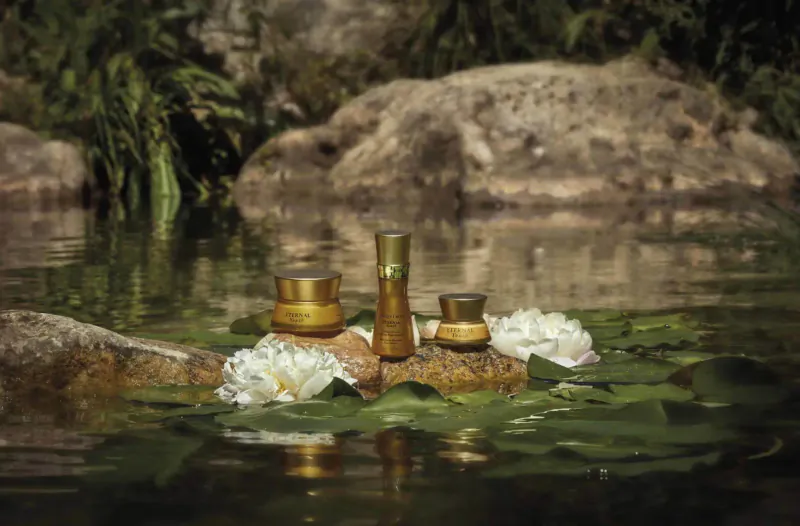 Gold skincare jars and bottle arranged with white lotus flowers on rocks in serene pond, lush greenery backdrop