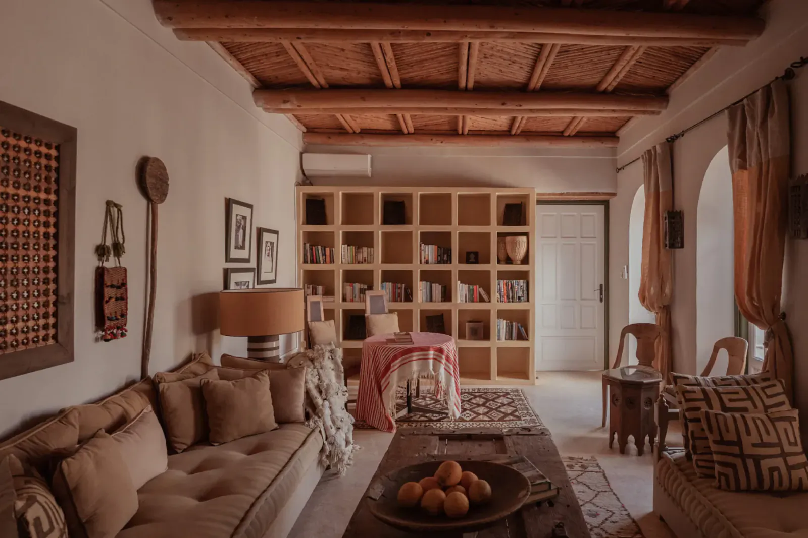 Cozy Moroccan villa living room with wooden beams, beige sofa, bookshelves, orange bowl on table, and woven decor.