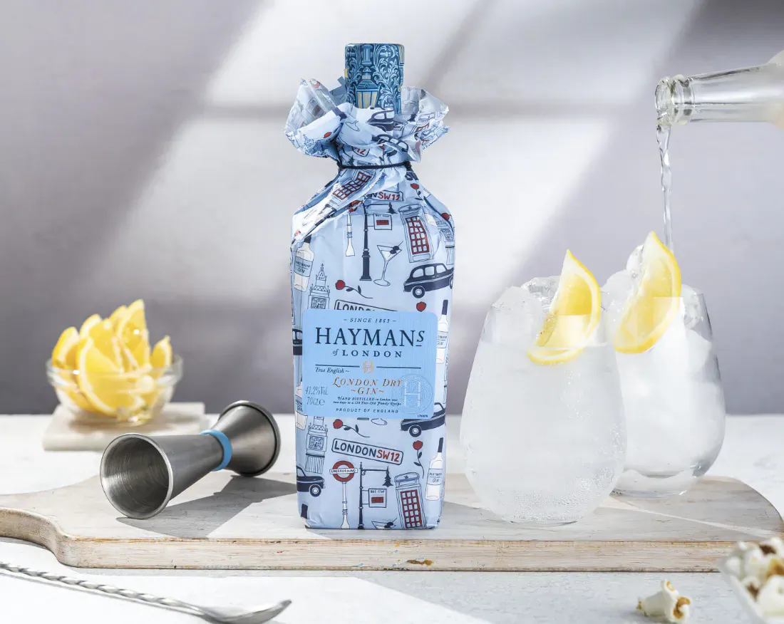 Hayman's London Dry Gin bottle in limited edition London wrap pouring into two glasses with lemon slices on a wooden board.