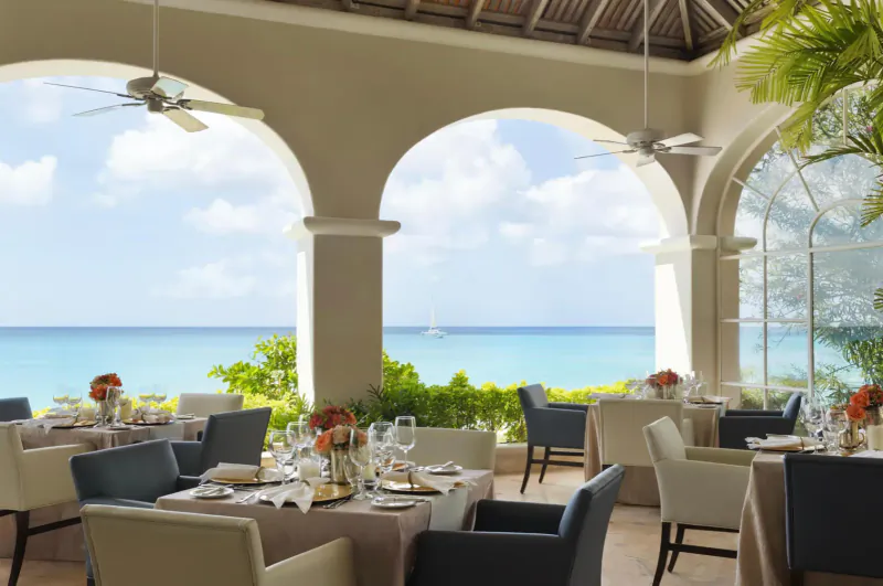 Elegant open-air dining pavilion at Fairmont Royal Pavilion with ocean view, arched windows, potted plants, and set tables with flowers.
