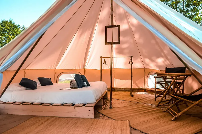 Spacious luxury glamping tent interior with king bed, pillows, desk, chair on wooden floor amid trees