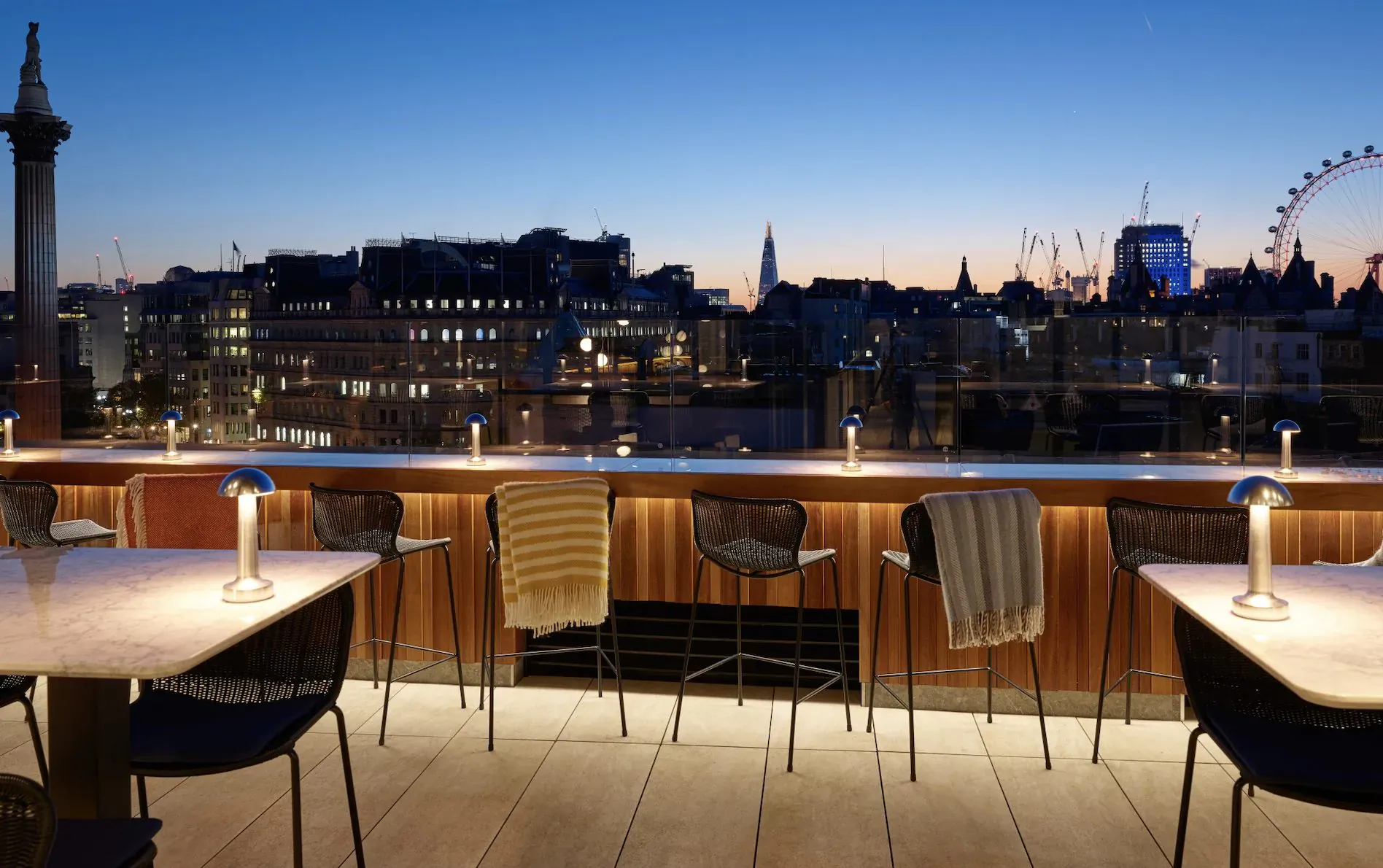 Rooftop bar at dusk with London Eye and skyline view, elegant tables, chairs, and lamps ready for NYE celebration.