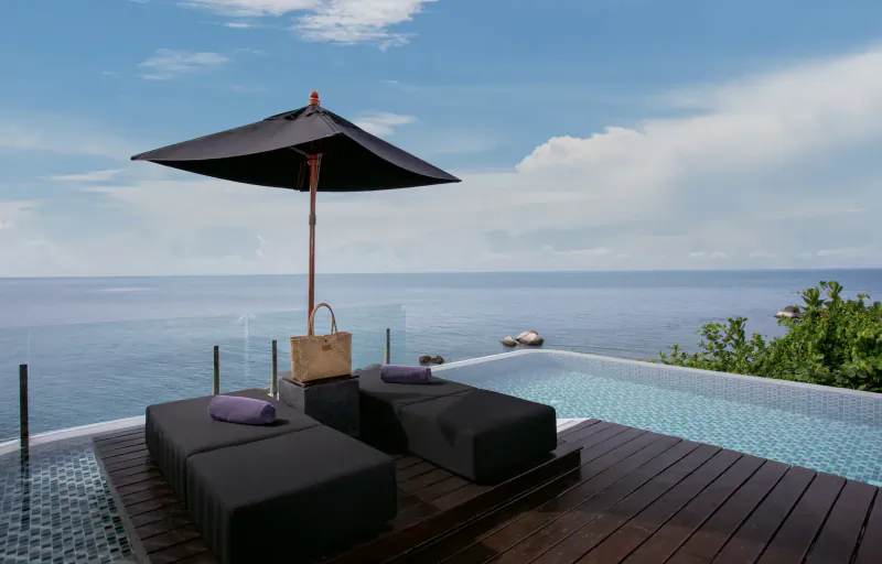 Infinity pool with black sun loungers, beige handbag, black umbrella on wooden deck overlooking ocean at Silavadee Pool Spa Resort