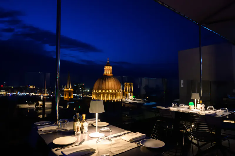 Twilight rooftop dining at Embassy Valletta Hotel with view of golden domes and city skyline, lit tables.