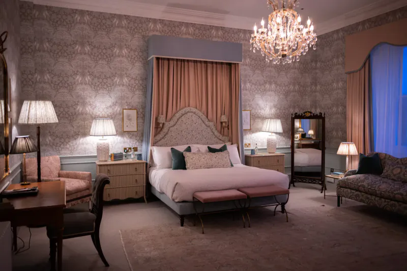 Elegant luxury hotel bedroom with ornate wallpaper, chandelier, canopied king bed, lamps, and velvet seating.