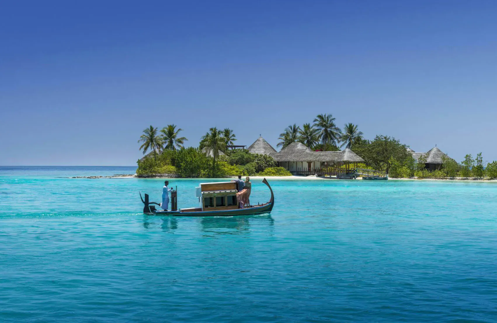 Traditional dhoni boat with two people glides on turquoise lagoon toward thatched-roof villas on lush Maldives island under blue sky.