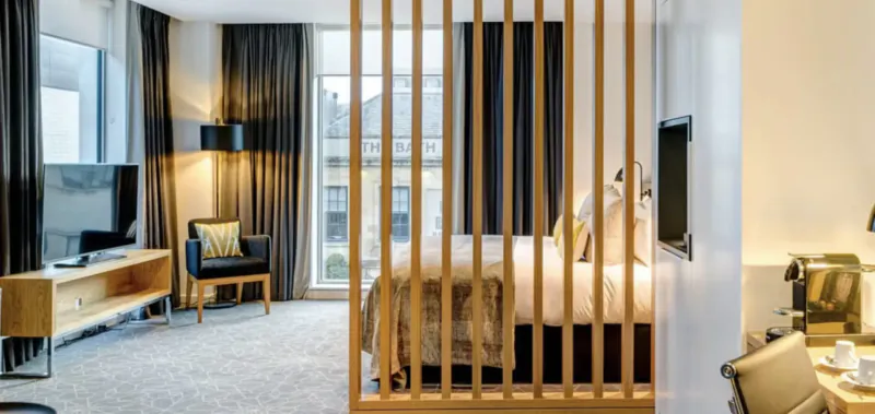 Modern hotel suite at Apex City of Bath with wooden partitioned king bed, TV, black chairs, lamps, and city view.