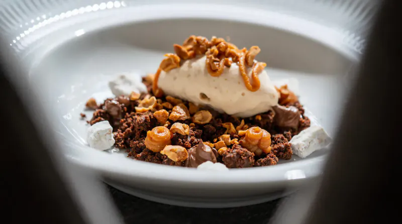 Close-up of dessert on white plate: chocolate crumble, peanut pieces, cream quenelle, and crispy topping