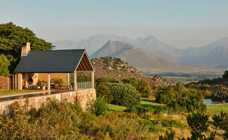 Stone wood-beamed gazebo on terrace overlooking Franschhoek mountains, lush gardens, and valley at sunset.