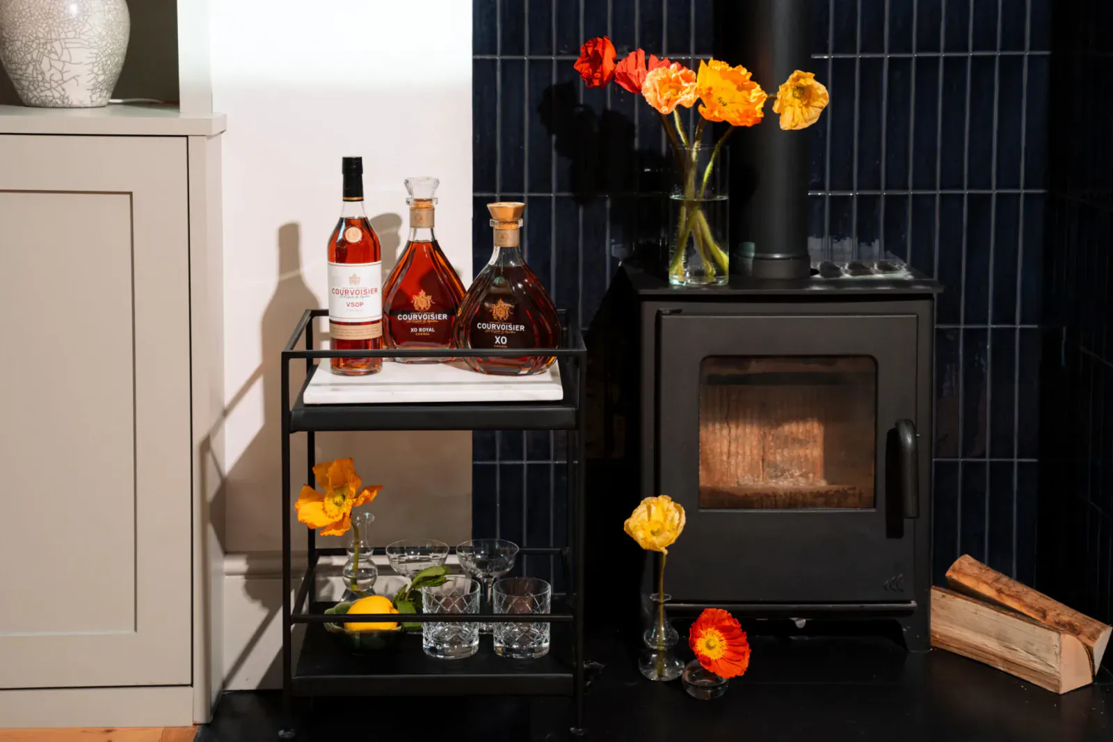 Elegant home bar cart with Cognac bottles, glasses, lemons, orange ranunculus flowers beside black wood stove on tiled wall.