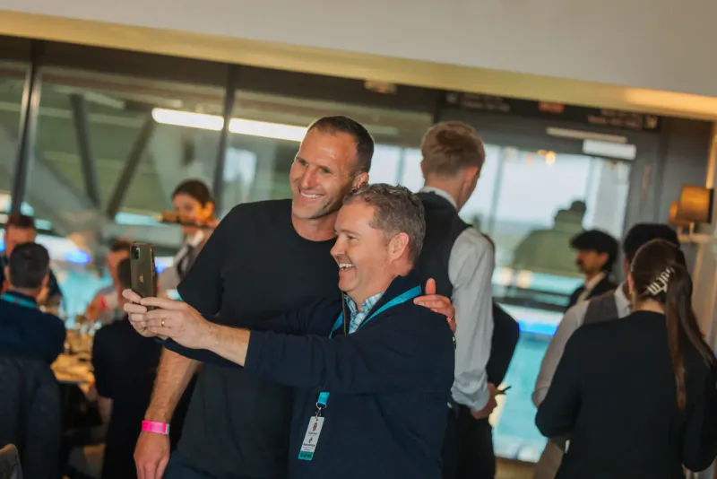Two smiling men in lanyards taking a selfie at rugby event, with staff and poolside crowd behind.