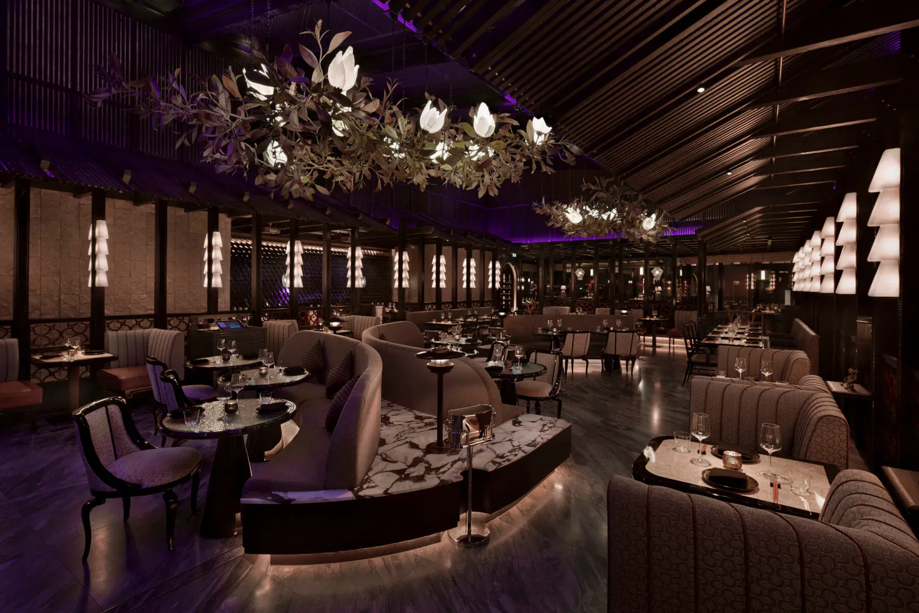 Elegant Tattu London lounge with curved velvet seating, marble tables, purple lighting, and hanging floral chandeliers