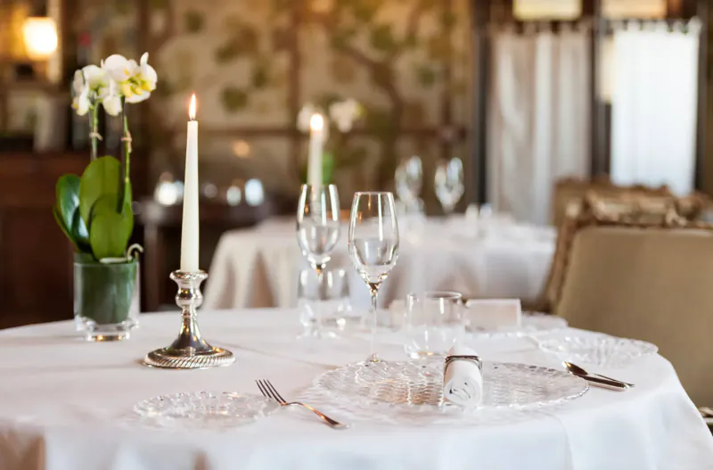 Elegant Tuscan dining table with white orchid, candles, wine glasses, salt shaker on white cloth, floral wallpaper backdrop.