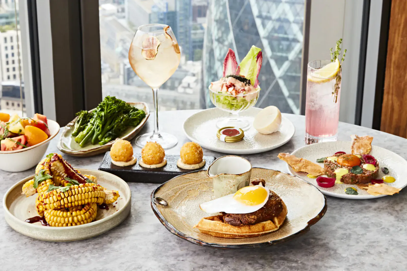 Duck and waffle dining spread with gin cocktails, salads, corn, and city skyline view from high window.