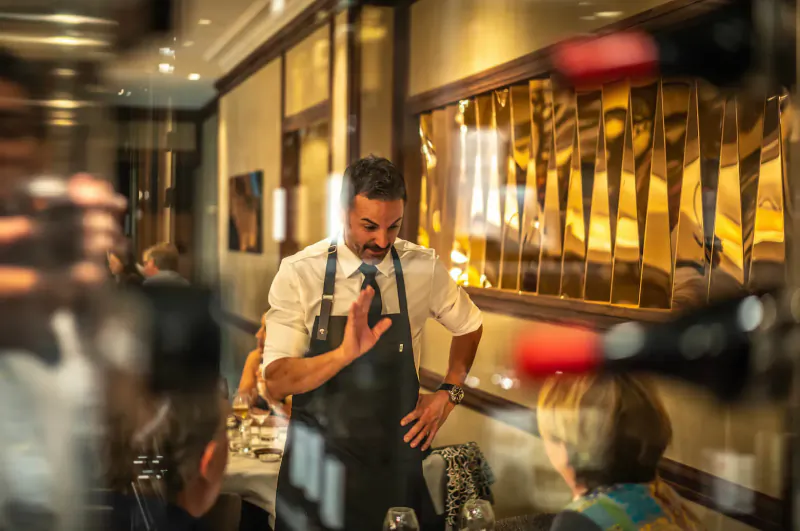 Bearded waiter in white shirt and black apron gestures welcomingly to diners beside wine bottles in elegant Michelin-starred restaurant.