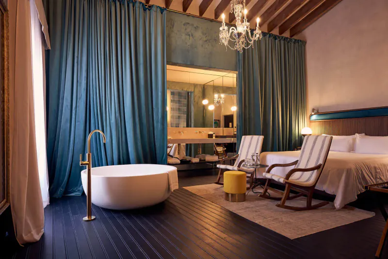 Elegant hotel suite with freestanding bathtub, wooden beams, teal curtains, chandelier, and rattan chairs.