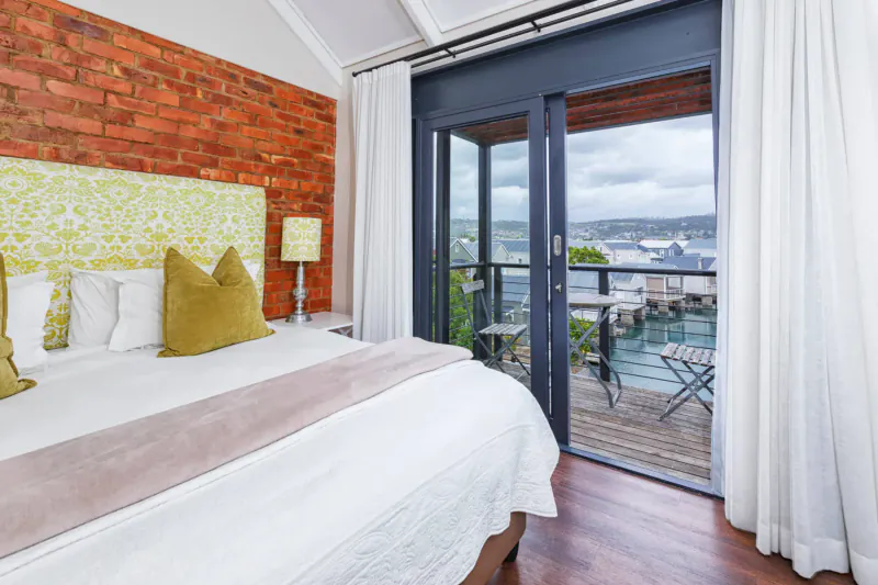 Luxury hotel bedroom with green pillows on bed, brick walls, open doors to balcony overlooking harbor marina.