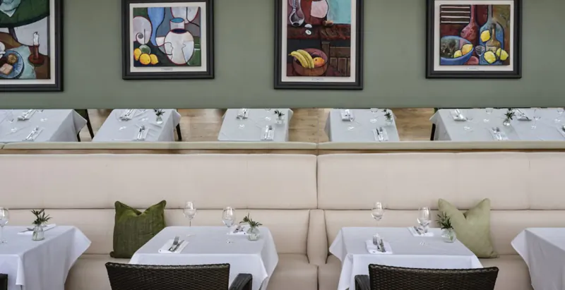 Elegant dining area at The Bristol Gallery with white tablecloths, cushioned booths, and colorful still life paintings of fruits and chef.