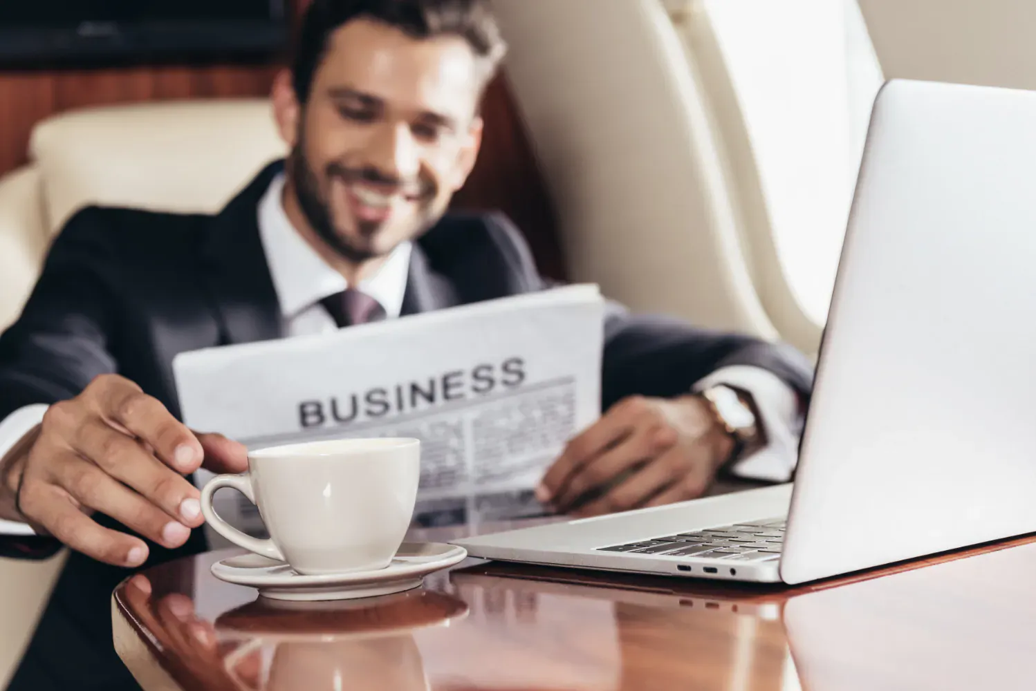 Smiling businessman in suit reading 'BUSINESS' newspaper, sipping coffee from white cup beside MacBook in luxury office.