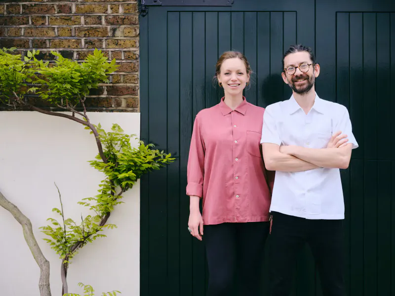 Smiling woman in pink shirt and man in white shirt with arms crossed stand in front of dark green garage door, brick wall, and green plant.
